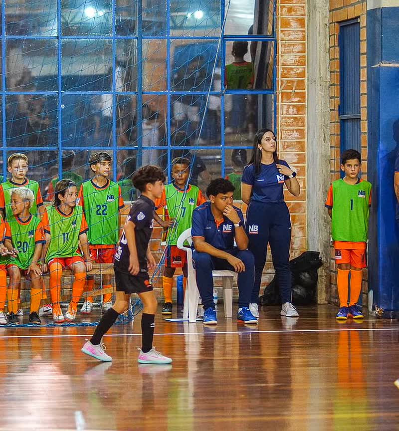 Bruna à beira da quadra acompanhando o banco de atletas da base durante um jogo de futsal