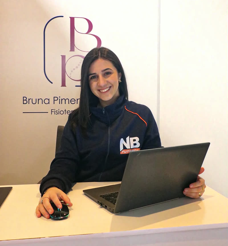 Bruna Pimentel Pahim, fisioterapeuta traumato-ortopédica e esportiva, sentada à mesa de trabalho com a marca dela ao fundo